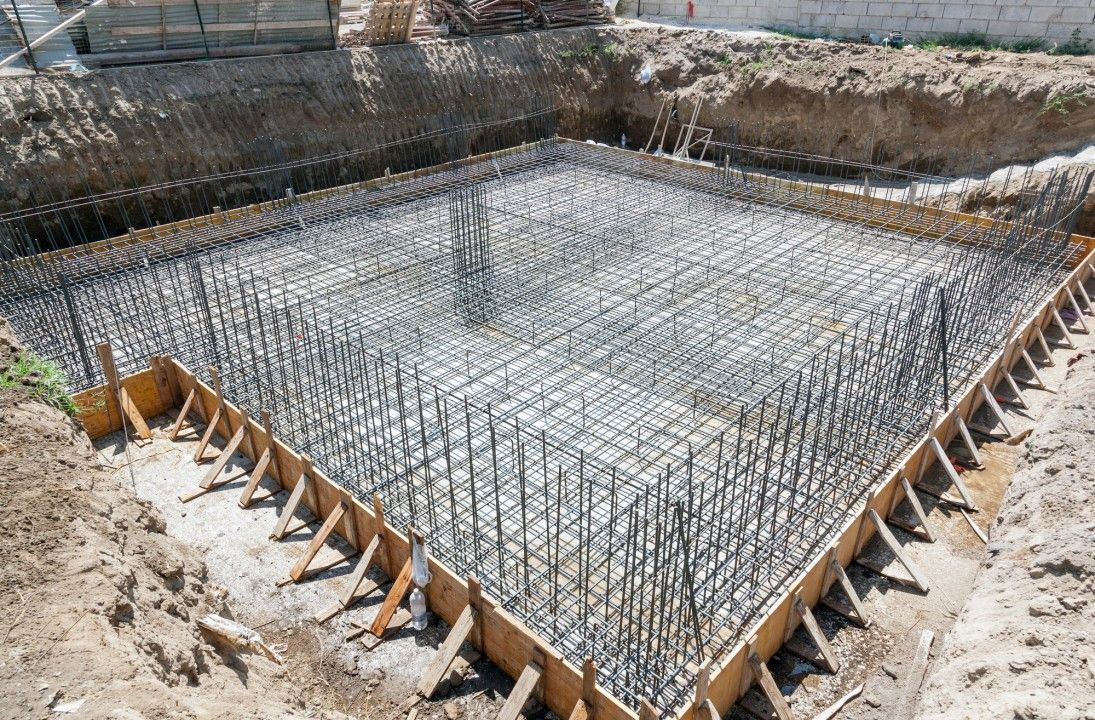 A grid of steel rebar reinforcement prepared inside a wooden formwork for a concrete foundation at a construction site.