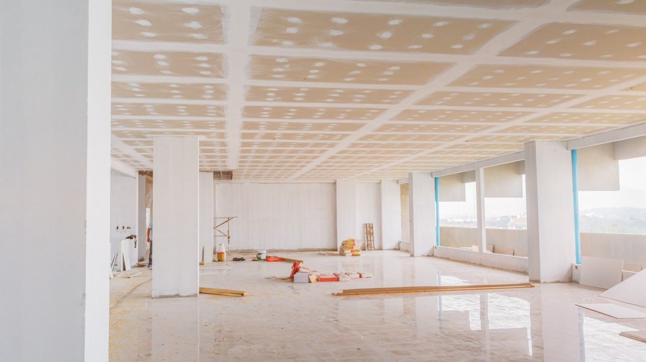 A spacious, unfinished interior room with a grid-patterned drywall ceiling, white concrete pillars, and a tiled floor.
