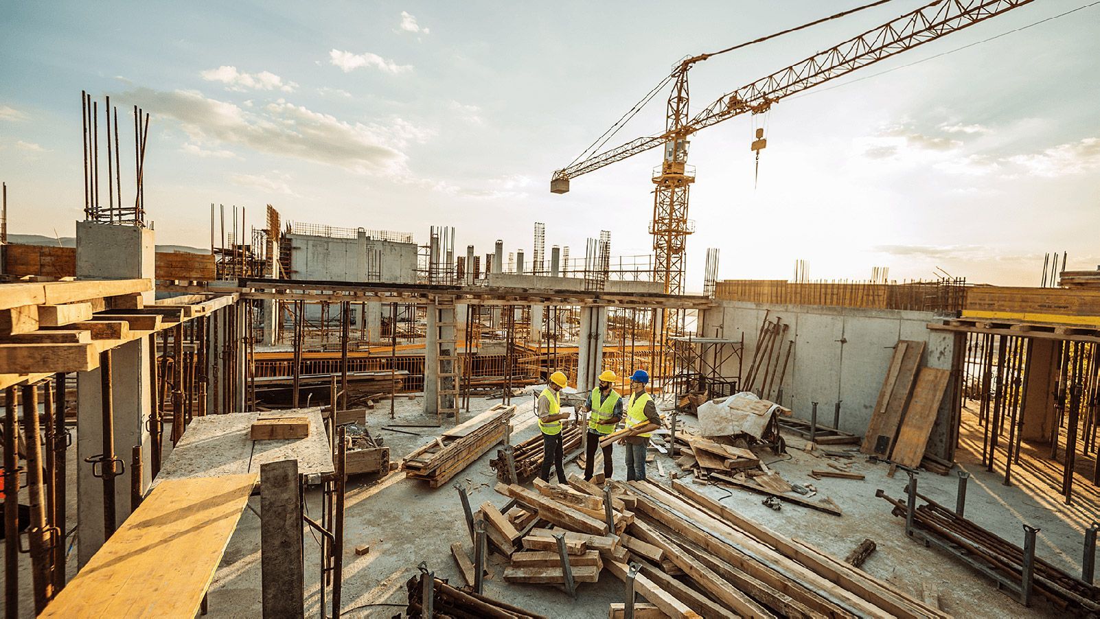 Three construction workers in hard hats stand on a building site at sunset, with a large crane visible in the background.