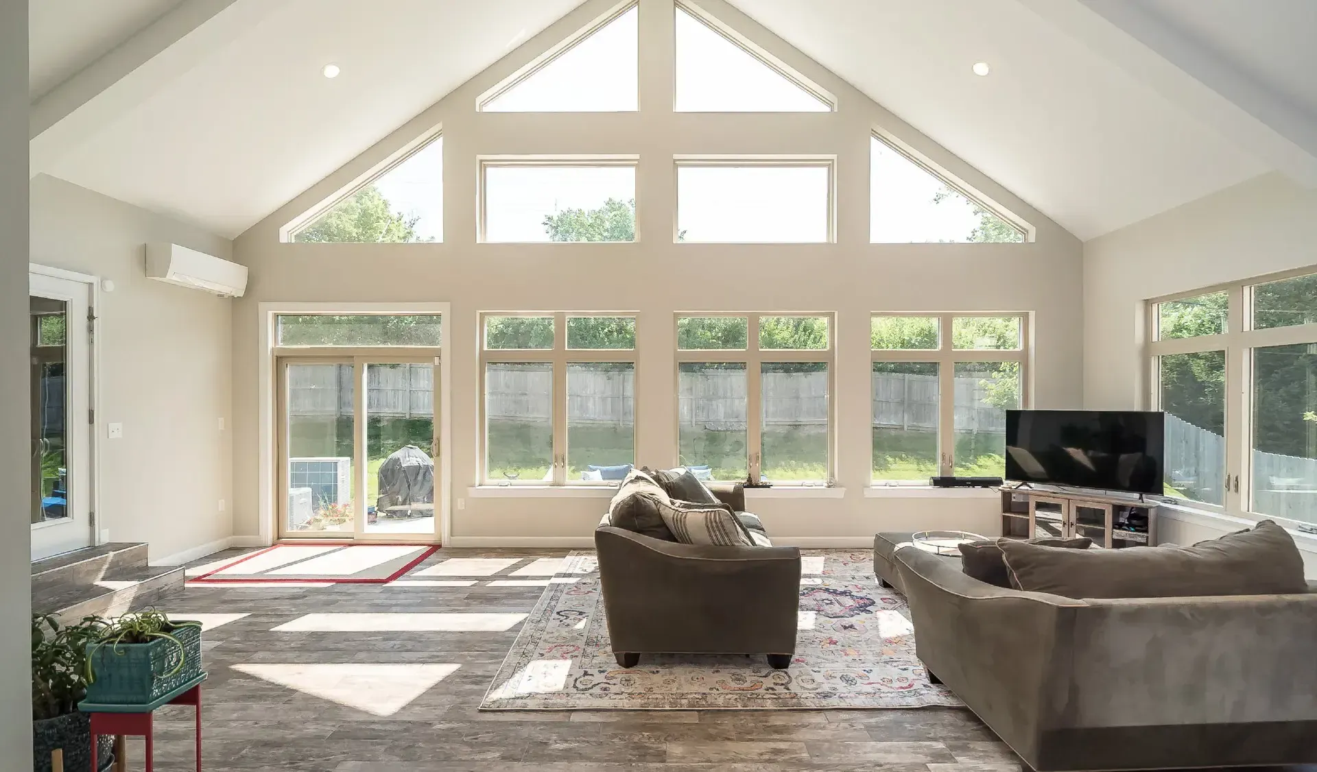 Spacious sunroom with vaulted ceilings, large windows, sliding glass door, and two grey couches facing a TV.