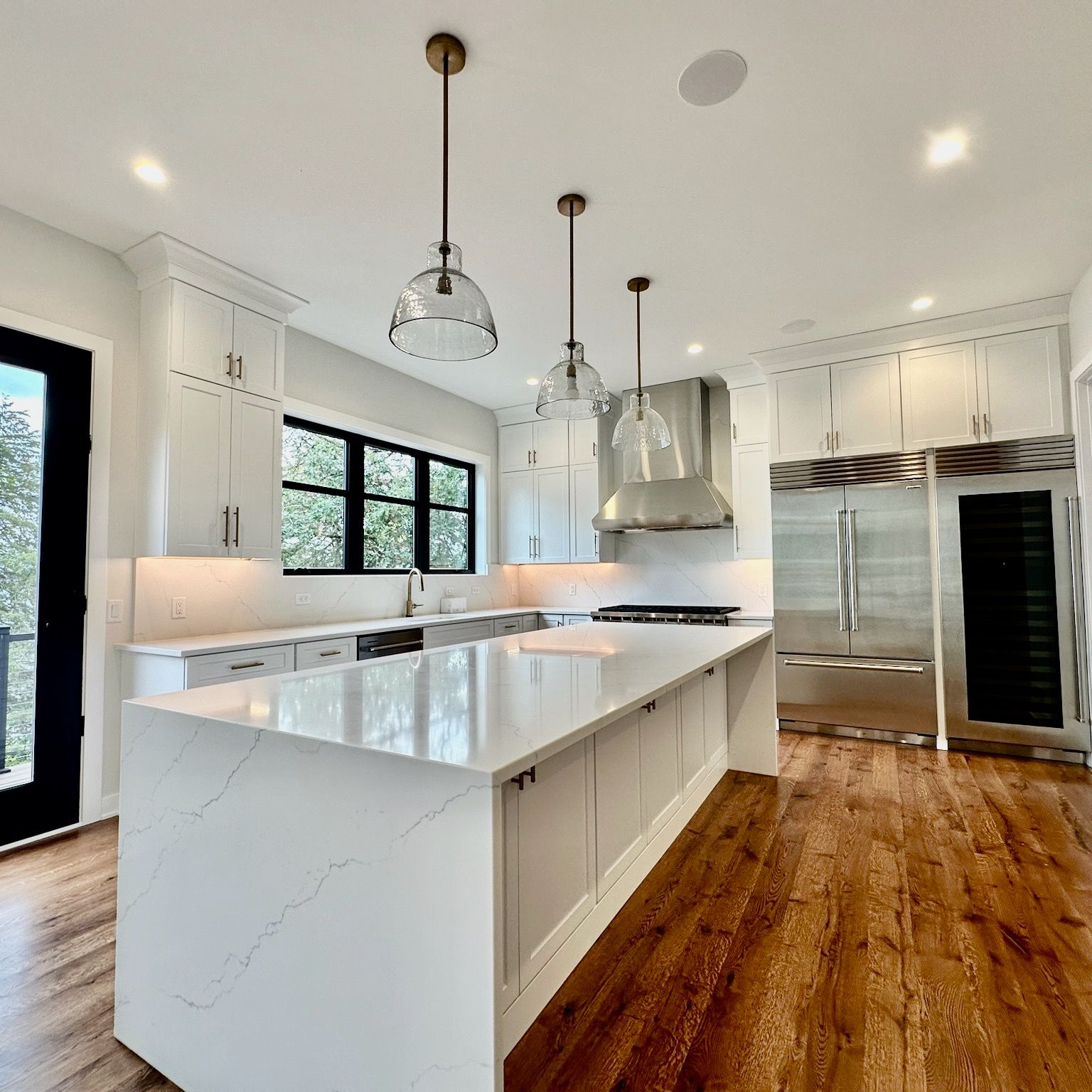 Modern white kitchen with a quartz island, wood flooring, stainless steel appliances, and three pendant lights.