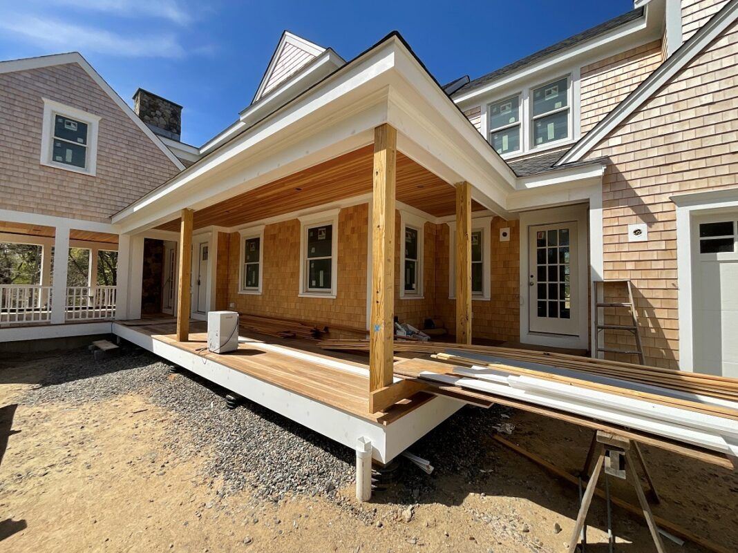 A new wooden porch structure under construction attached to a shingle-sided house with a gravel yard.