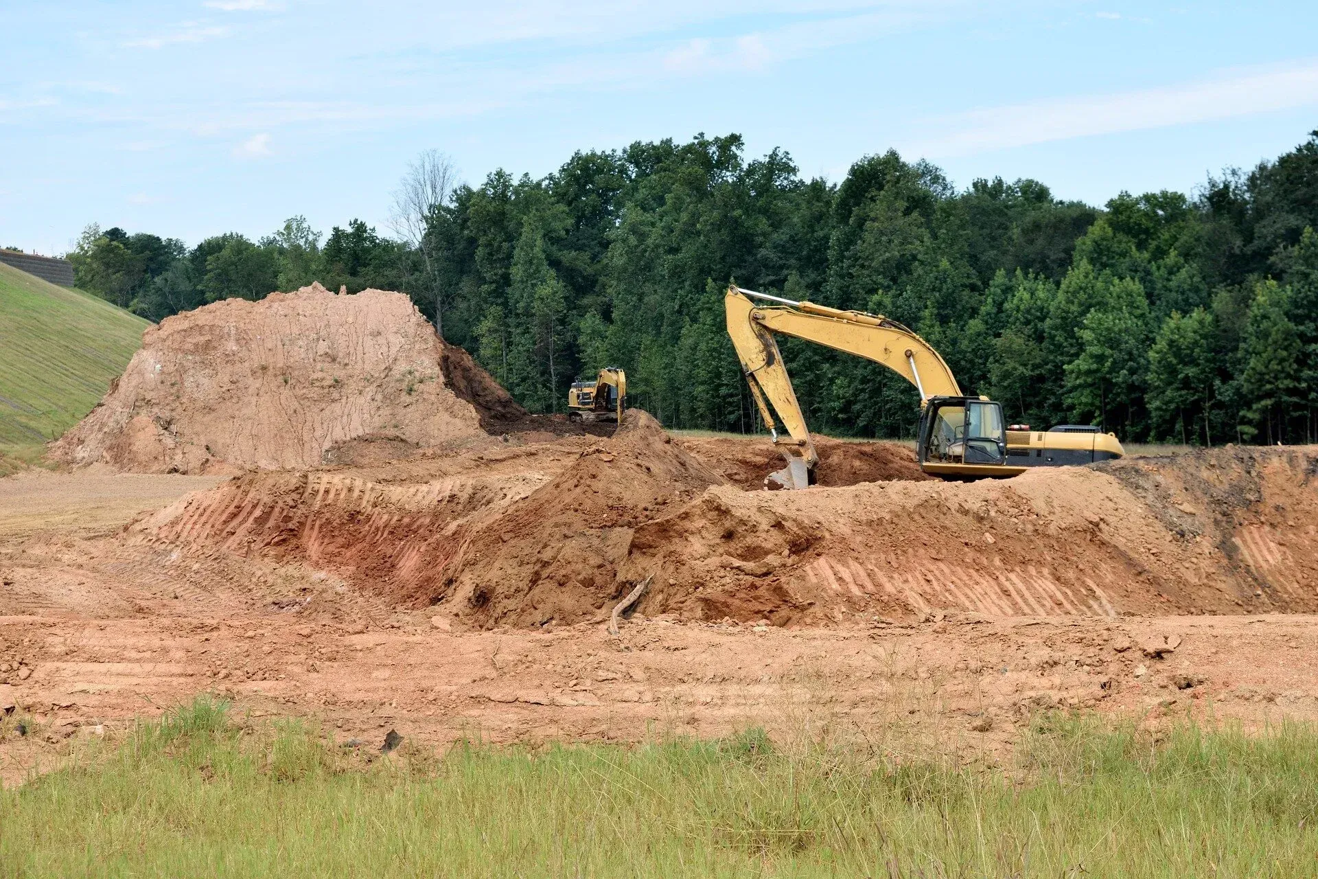 A yellow excavator works on a construction site with piles of excavated dirt in front of a dense treeline.