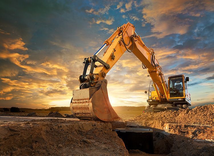 A yellow excavator sits in a dirt pit under a dramatic sunset sky.