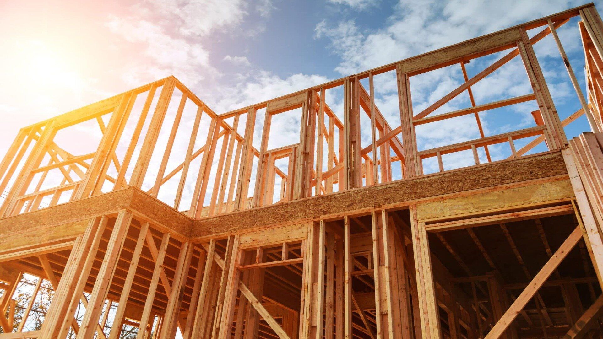 The wooden frame of a two-story house under construction against a bright sky with sunlight streaming from the left.