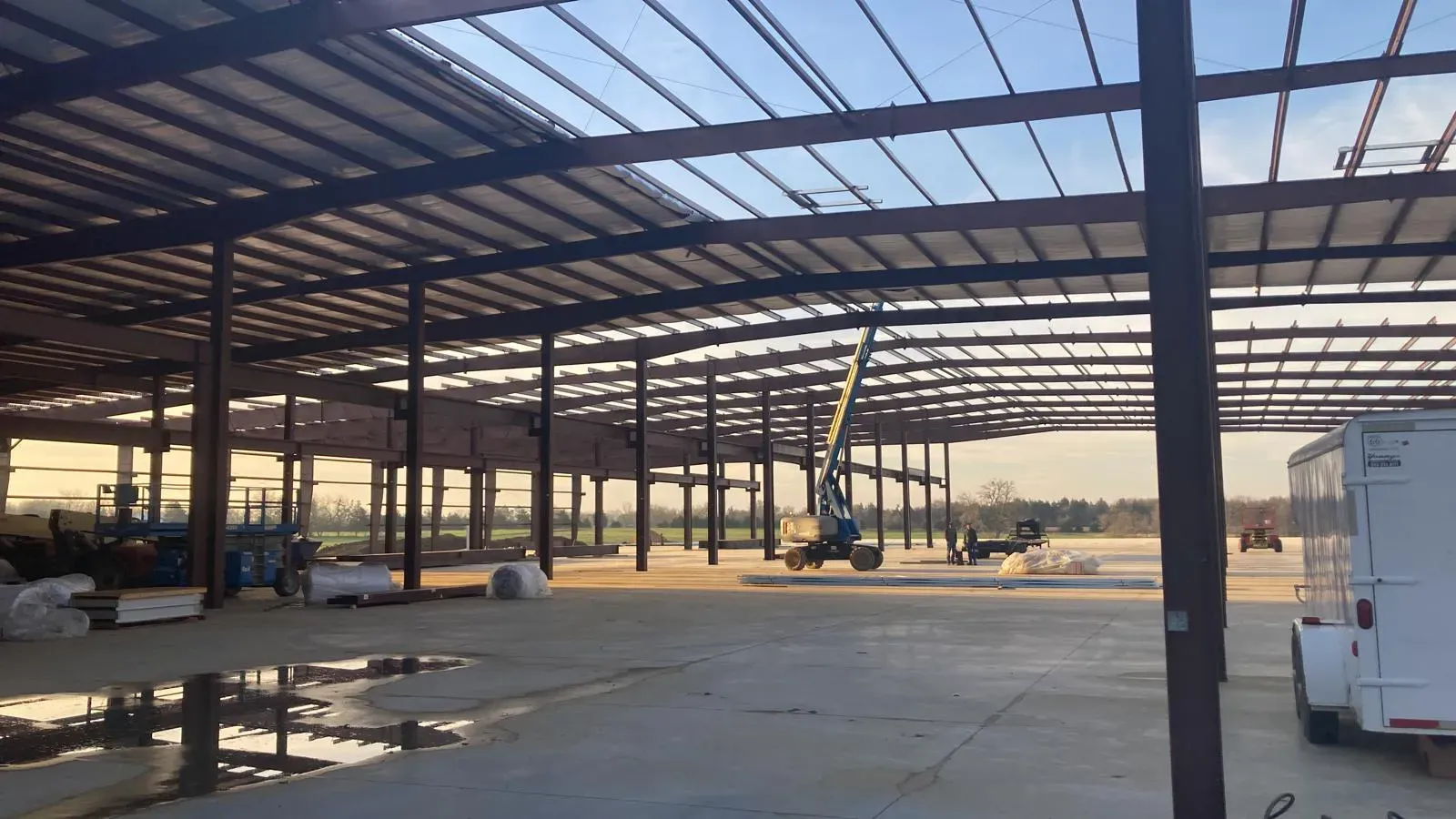 Large open construction site of a warehouse with a steel frame roof, support beams, and equipment under a clear sky.