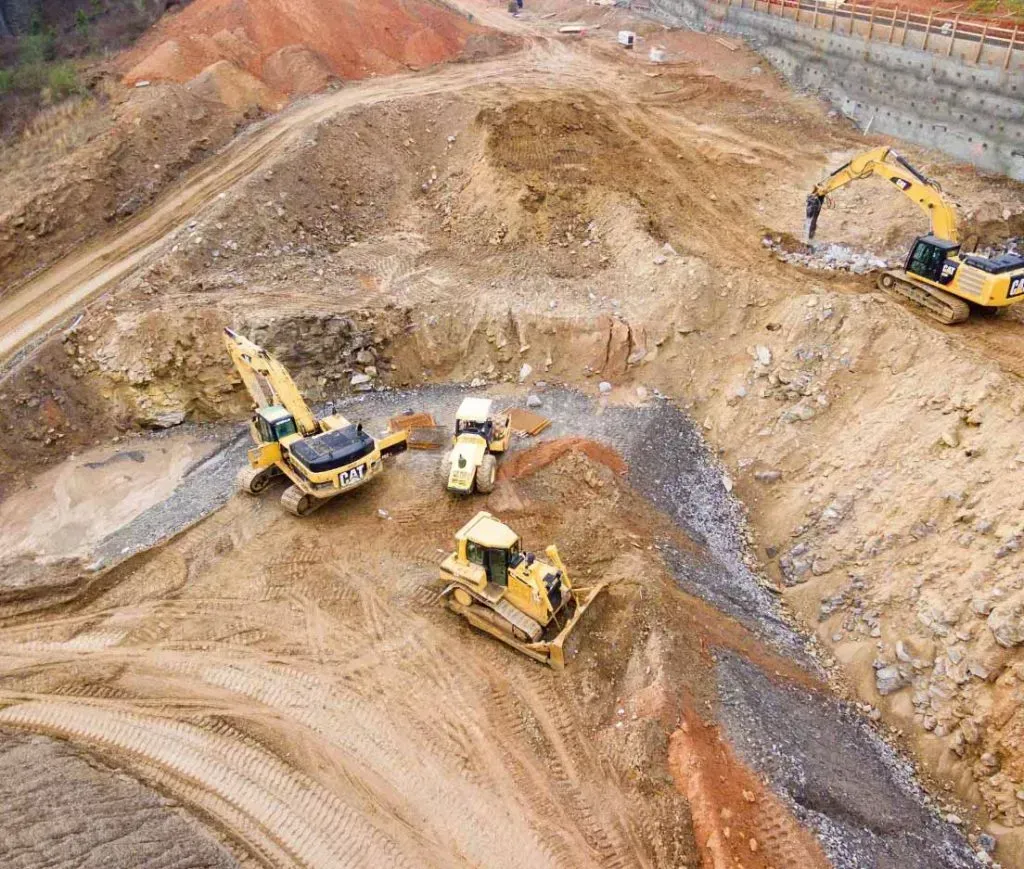 Aerial view of construction site with three yellow excavators and a bulldozer working on uneven dirt terrain.