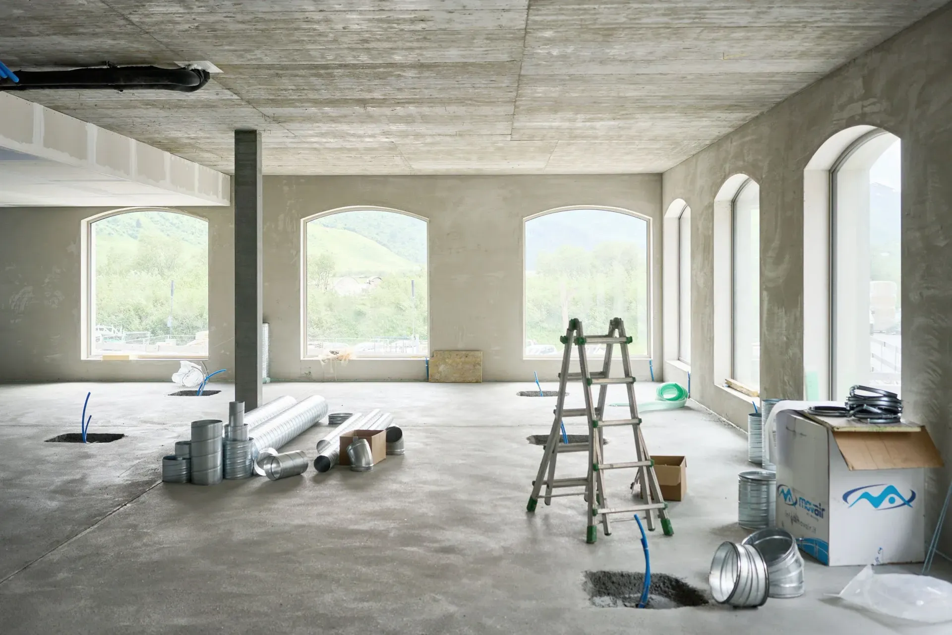 Unfinished interior construction site with raw concrete walls, large arched windows, a ladder, and scattered materials.