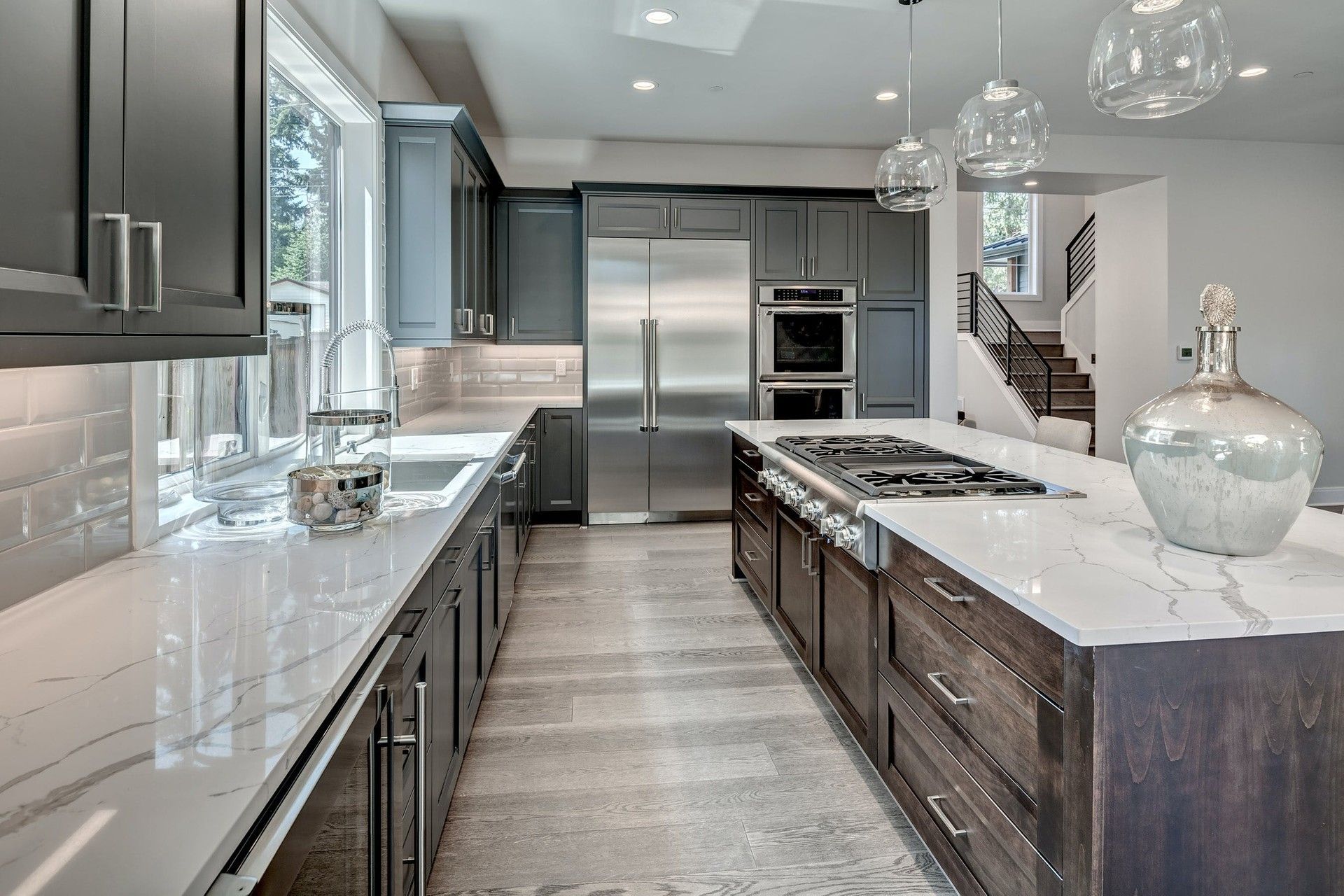 Modern kitchen featuring a large marble-topped island, stainless steel appliances, dark cabinetry, and wood flooring.