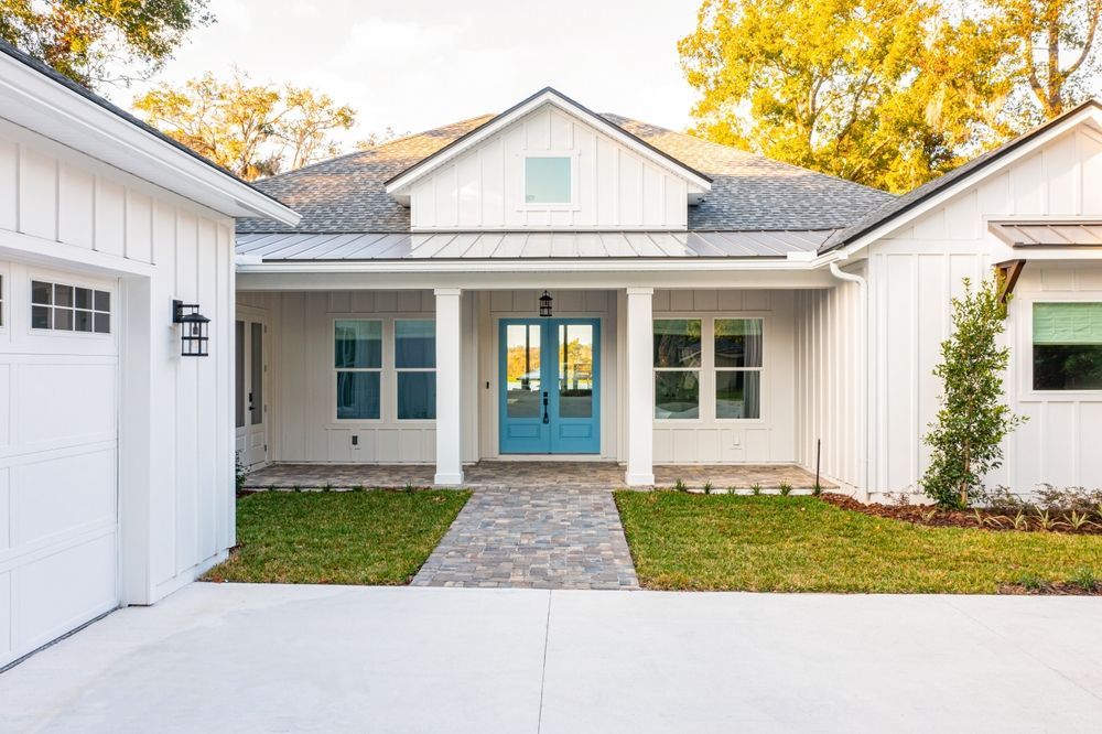 A modern white farmhouse with vertical board-and-batten siding, a gray shingle roof, and bright blue double front doors.