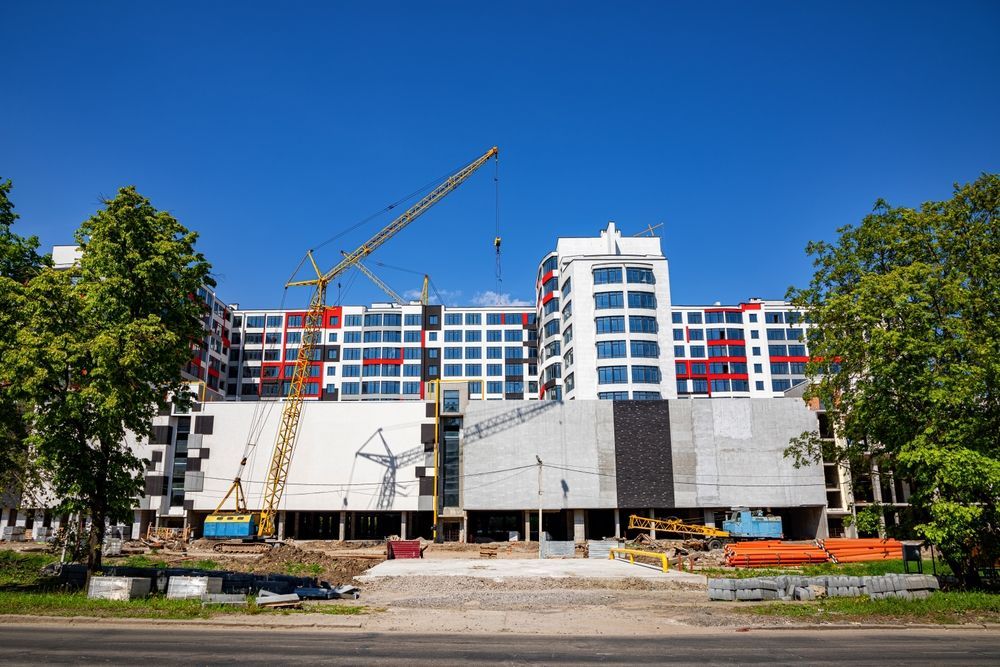 A large, partially constructed white apartment building under a clear blue sky, with a yellow crane on a dirt lot.