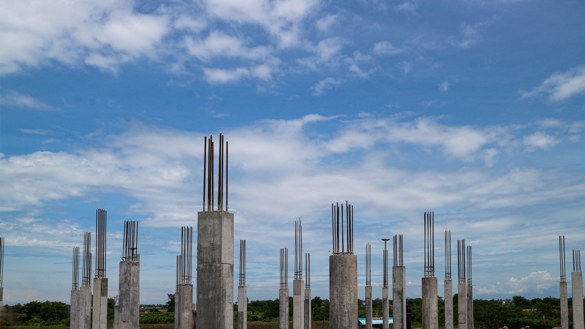 Concrete building columns with exposed steel rebar stand in an open field against a bright blue, cloudy sky.