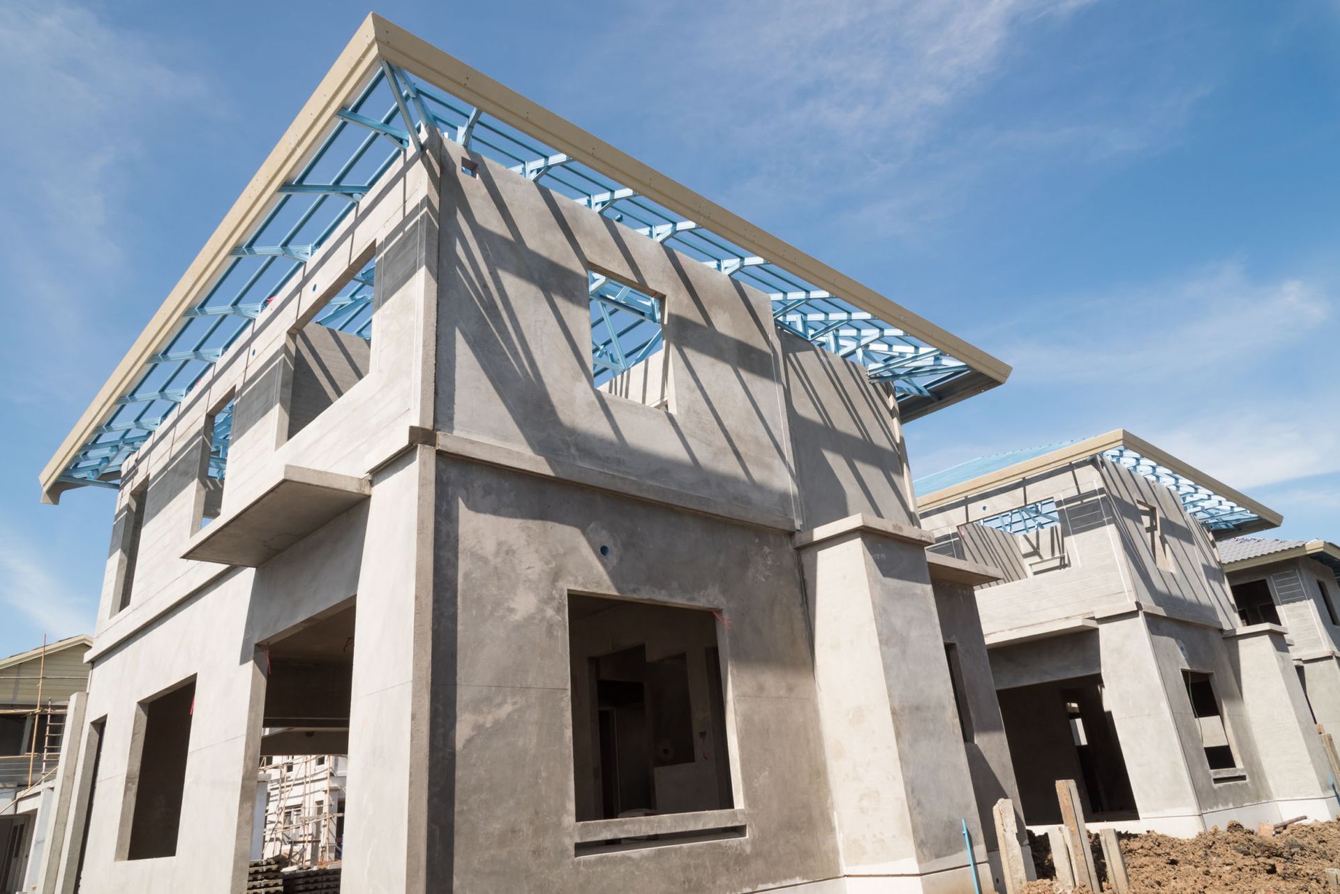Two unfinished concrete houses under a clear blue sky, showing exposed metal roof framing and unglazed window openings.