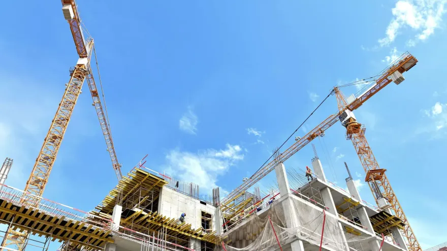Two tower cranes rise above a concrete building structure under a clear blue sky.