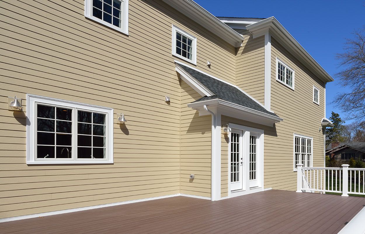 A beige, two-story house exterior with white trim, a brown deck, and a glass-paneled double door under a small roof.