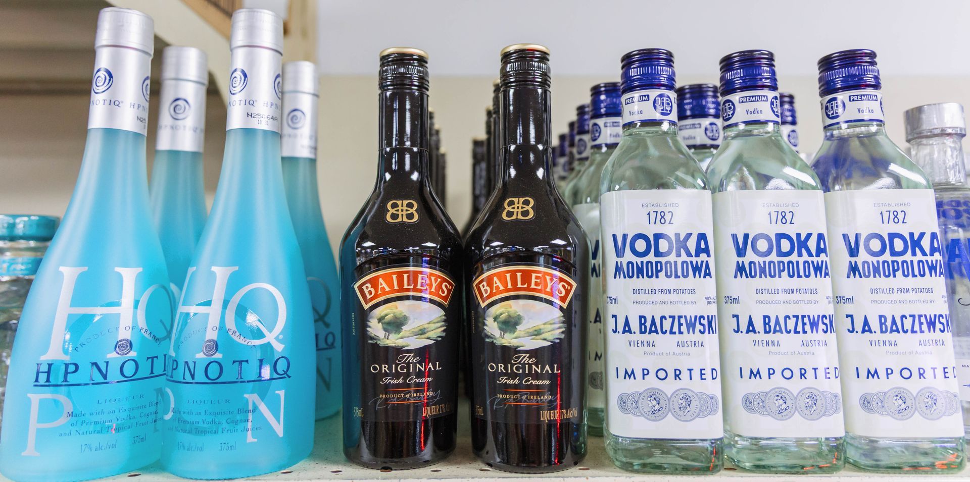 Bottles of various alcoholic beverages, including blue Hypnotiq, Bailey's Irish Cream, and vodka, on a shelf.