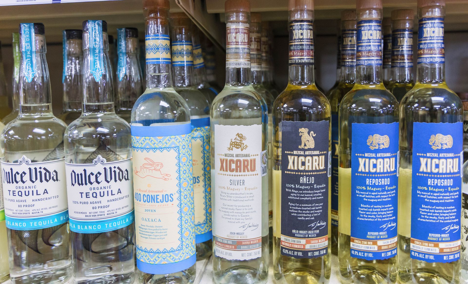 Bottles of tequila on a shelf; various labels and colors, mainly blue, brown, and clear glass.