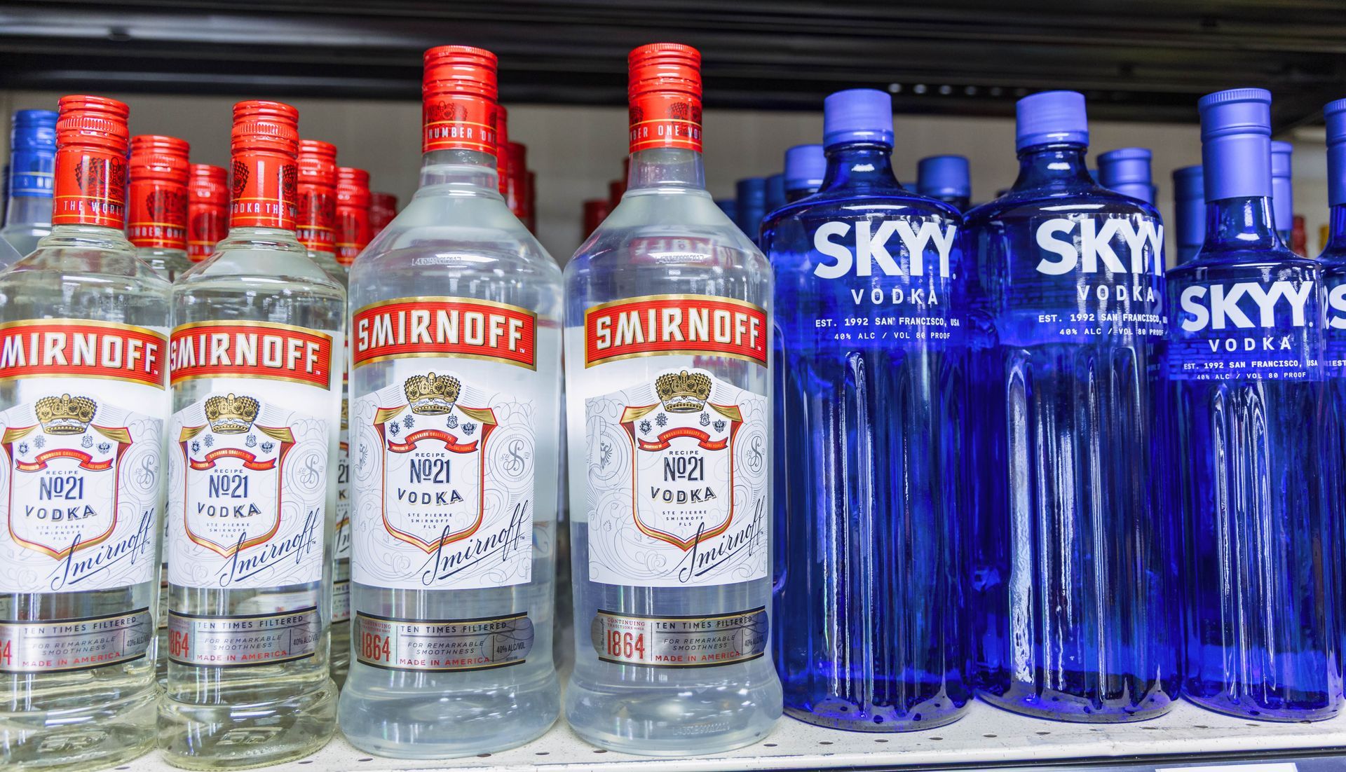 Bottles of Smirnoff and Skyy vodka on a store shelf, with red and blue bottle caps.