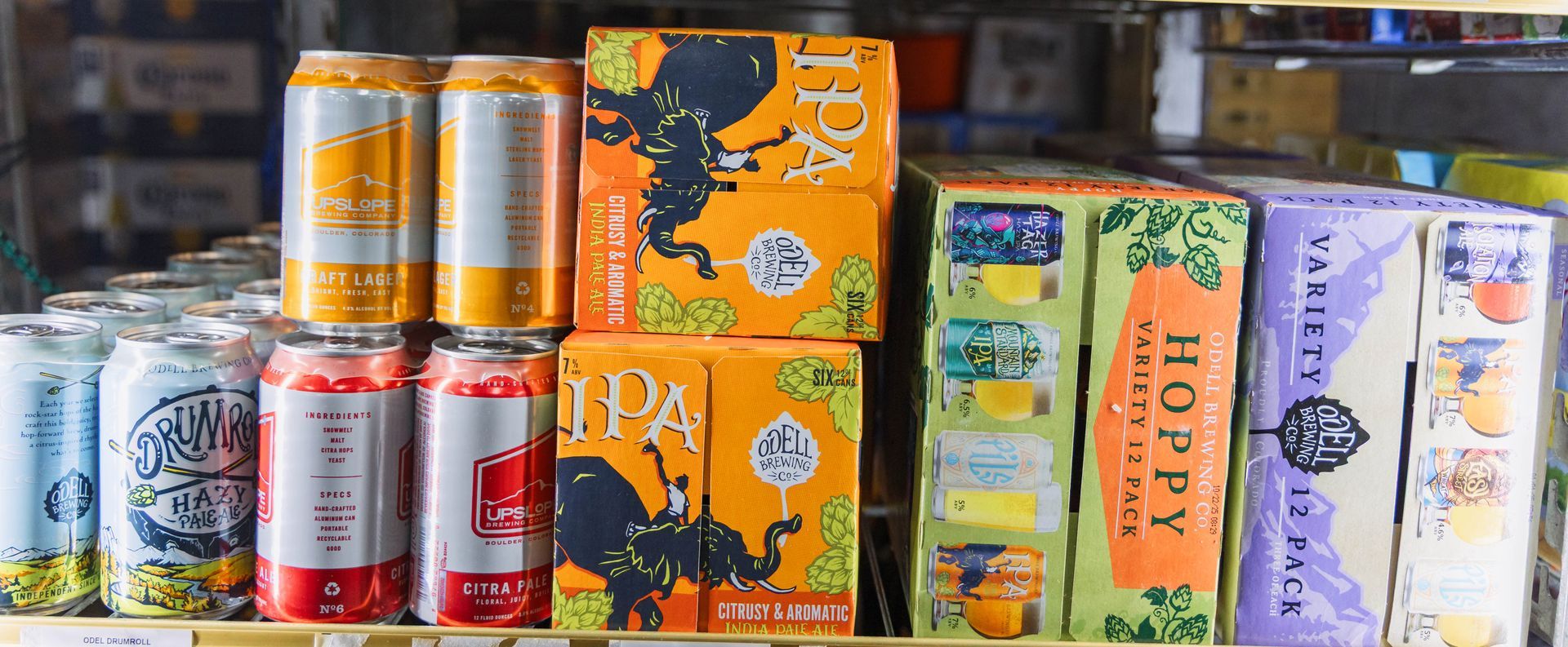 Shelves stocked with beer cans and boxes, including IPA and hoppy varieties, in a cooler.