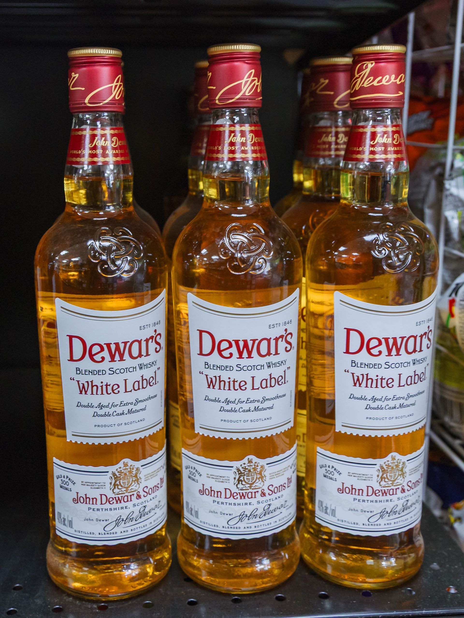Six bottles of Dewar's White Label scotch whiskey with gold labels and red caps, on a shelf.
