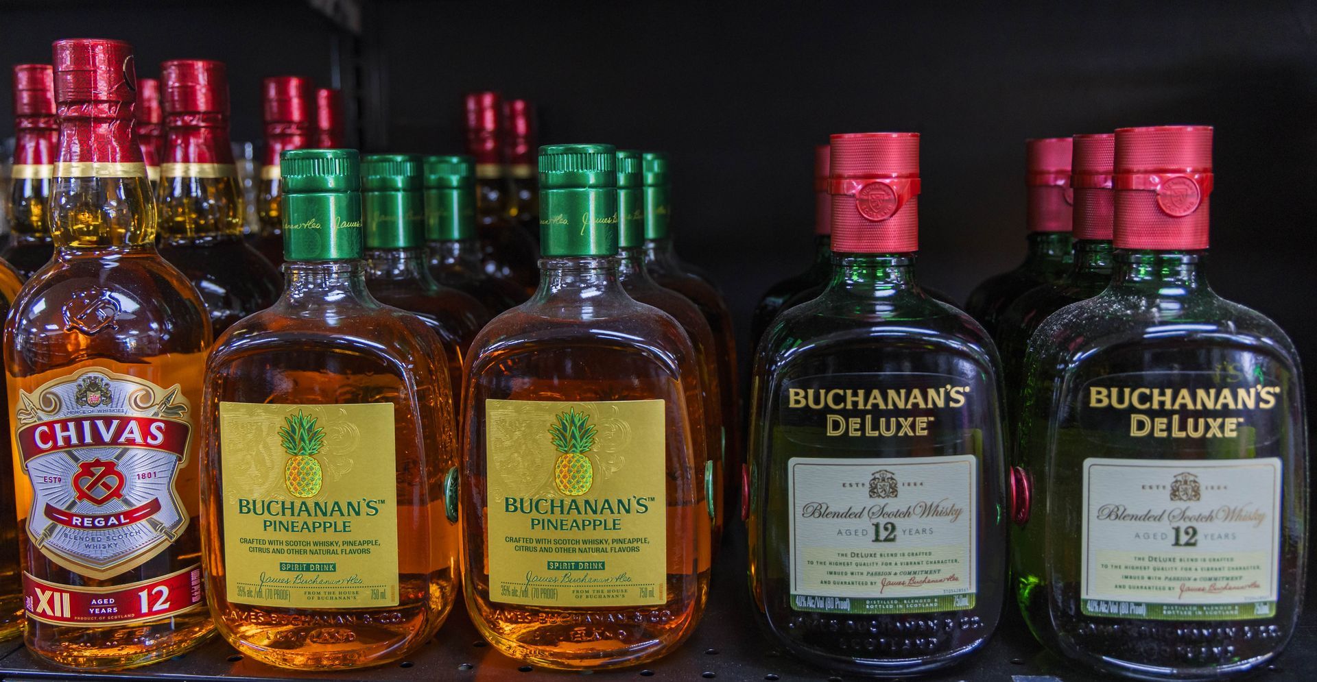 Bottles of Chivas Regal, Buchanan's, and Old Parr whiskey with red and green caps on a shelf.