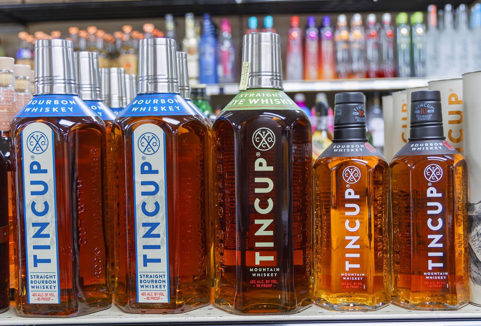 Bottles of TINCUP whiskey on a shelf, with a variety of labels and colors against a backdrop of other liquor bottles.