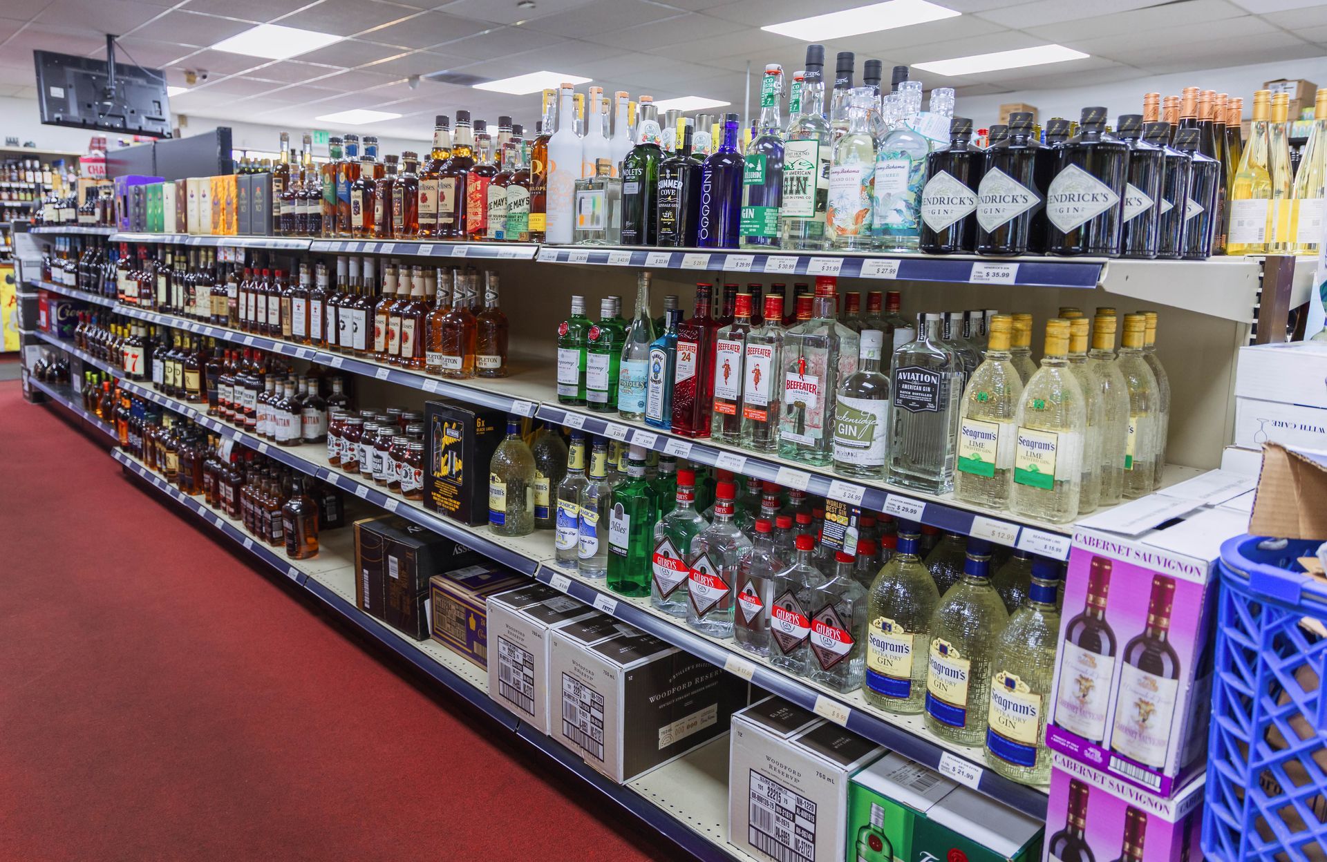 Shelves stocked with liquor bottles in a liquor store, red carpeted floor, overhead fluorescent lighting.