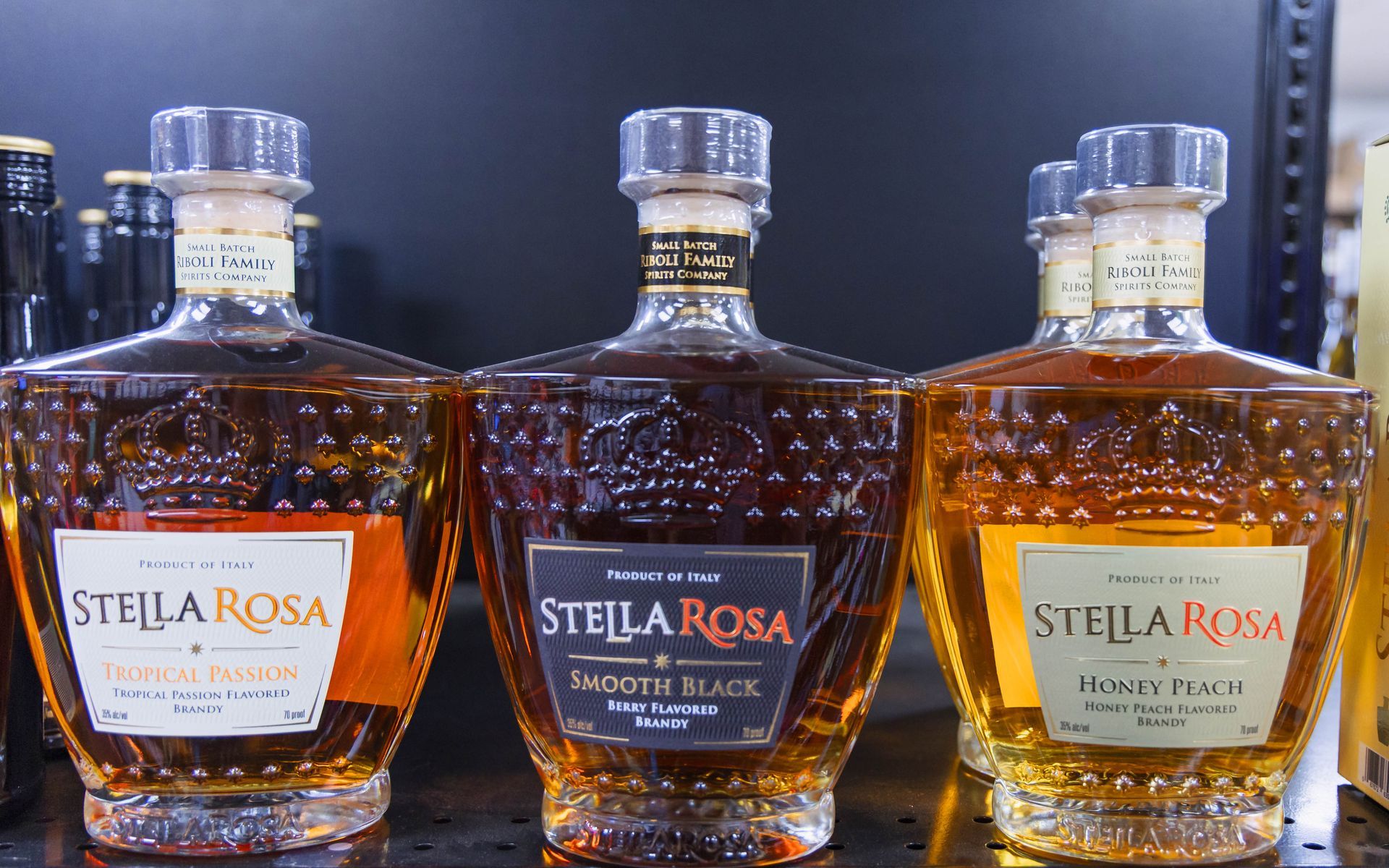 Three Stella Rosa wine bottles on a shelf: Original, Black, and Honey Peach, brown liquid, cork stoppers, and labels.