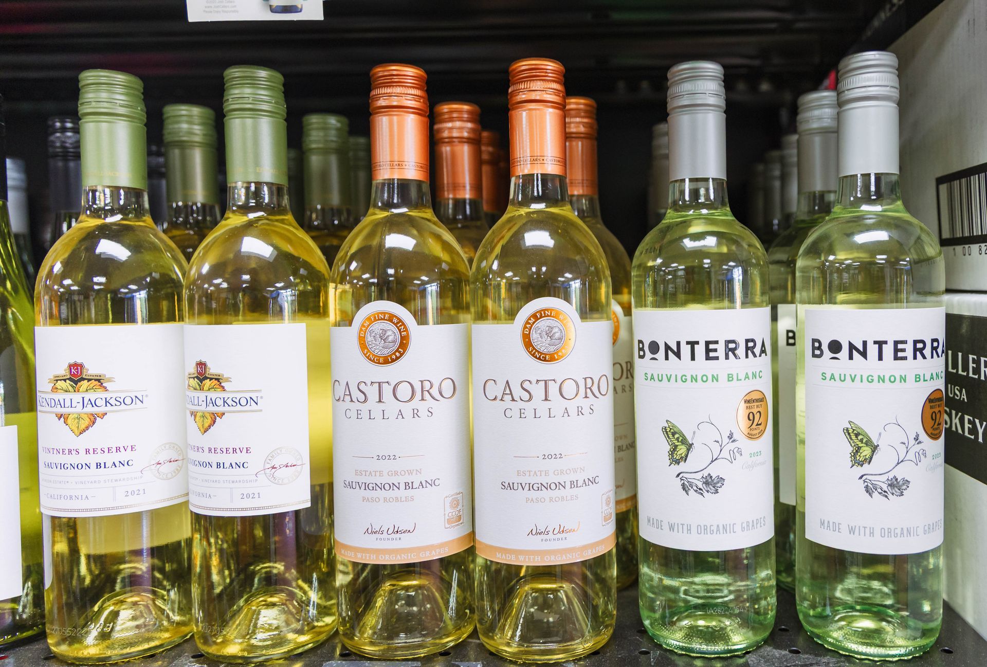 Wine bottles on a shelf, with white labels and a variety of colored bottle tops.