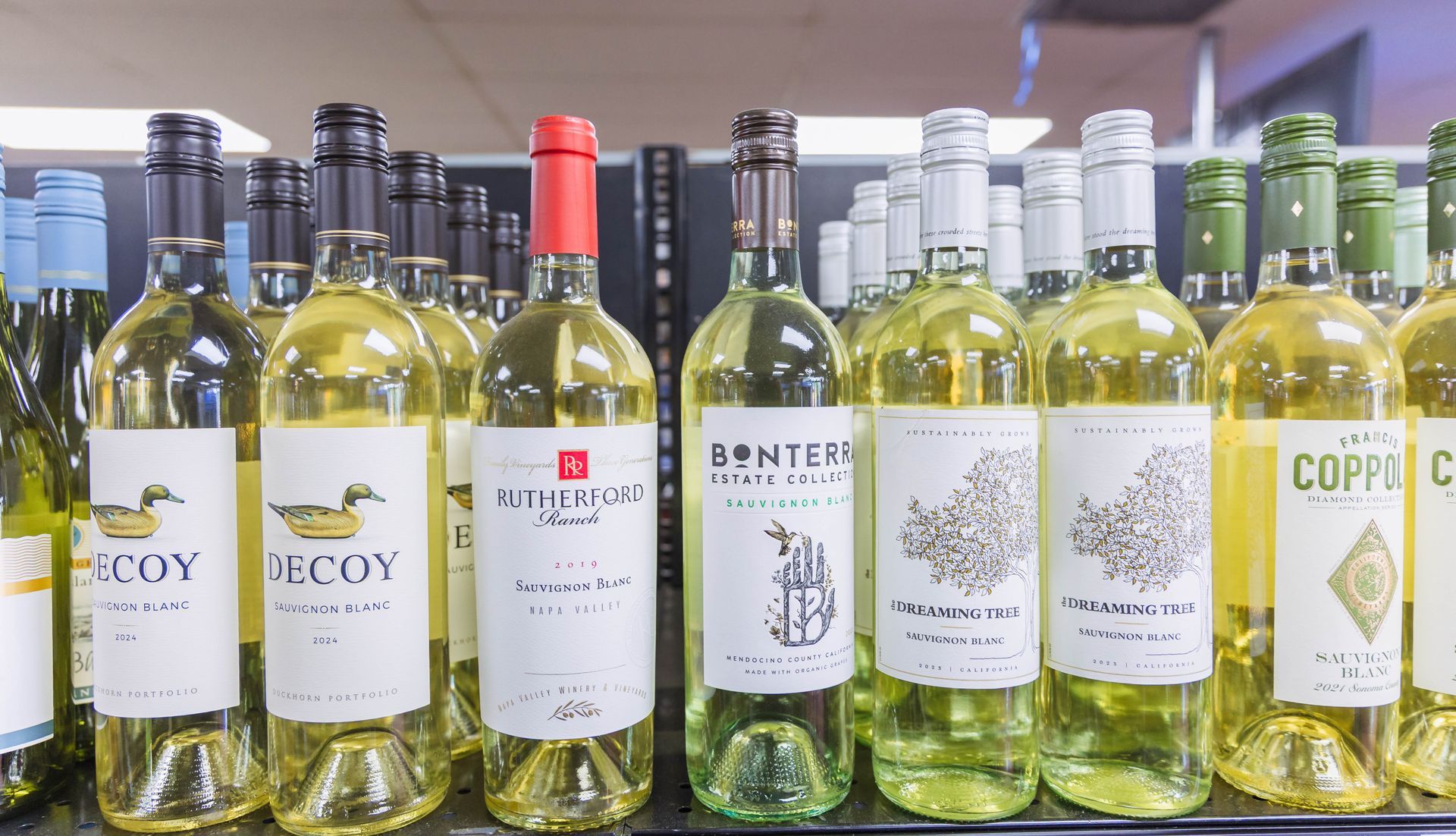 Bottles of white wine on a store shelf; various brands with light-colored labels, mostly green and white.