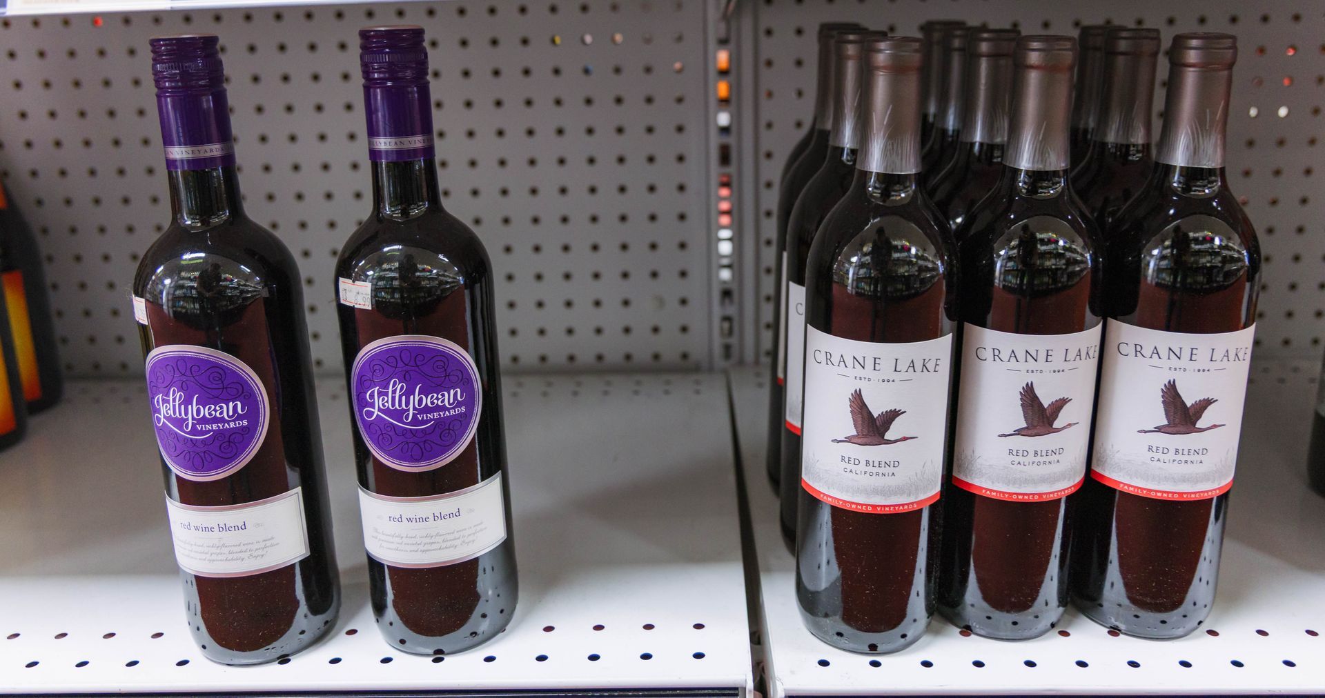 Two rows of wine bottles on a shelf, bottles with purple and white labels, and others with tan and white labels.