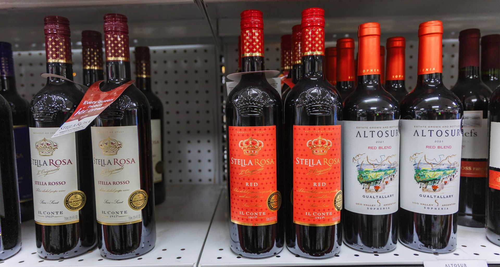 Wine bottles on a shelf, labels vary. Mostly dark glass, red and orange foil and labels.