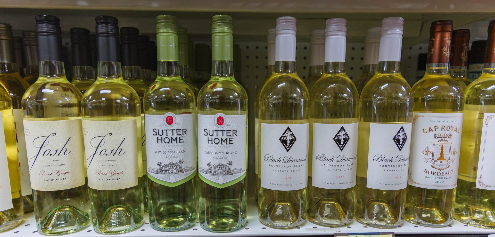 Wine bottles lined up on a store shelf. Various brands and colors, with white and green labels.