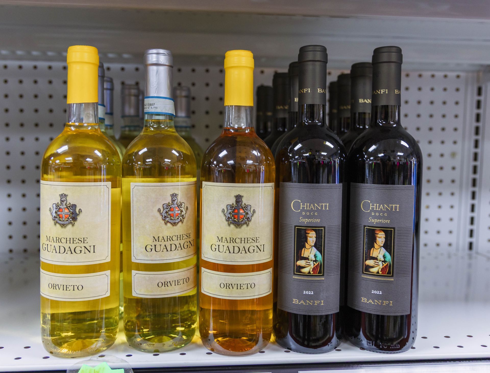 Bottles of wine on a shelf: yellow, amber and dark red, with labels, and a yellow and silver caps.