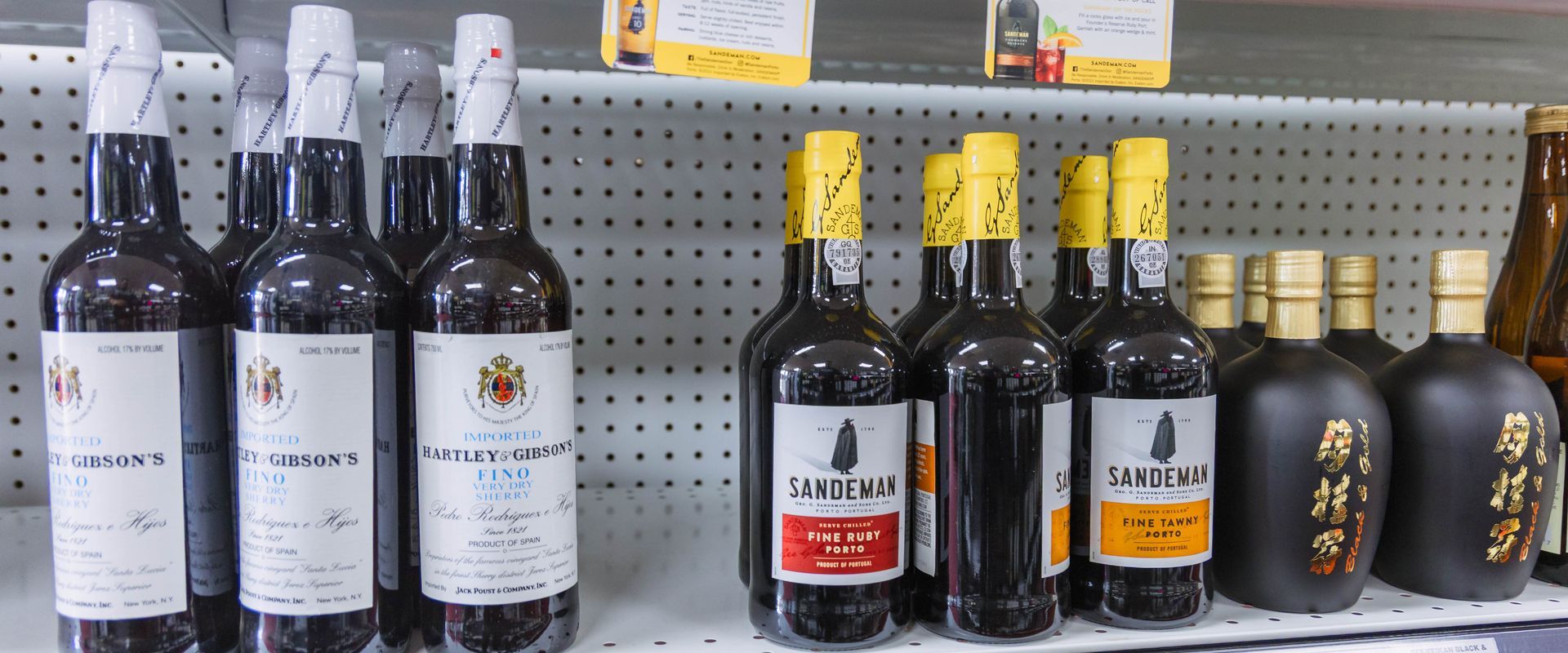 Bottles of various liquors on store shelves. Several have white labels and foil-wrapped tops. Dark glass bottles.