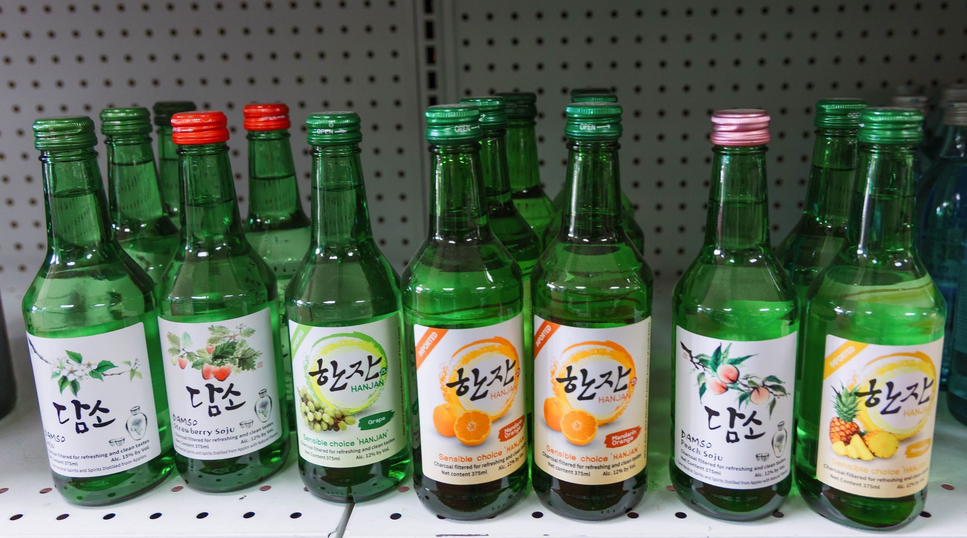 Bottles of soju on a shelf, in a variety of fruit flavors. Green bottles, colorful labels, and red and green bottle caps.