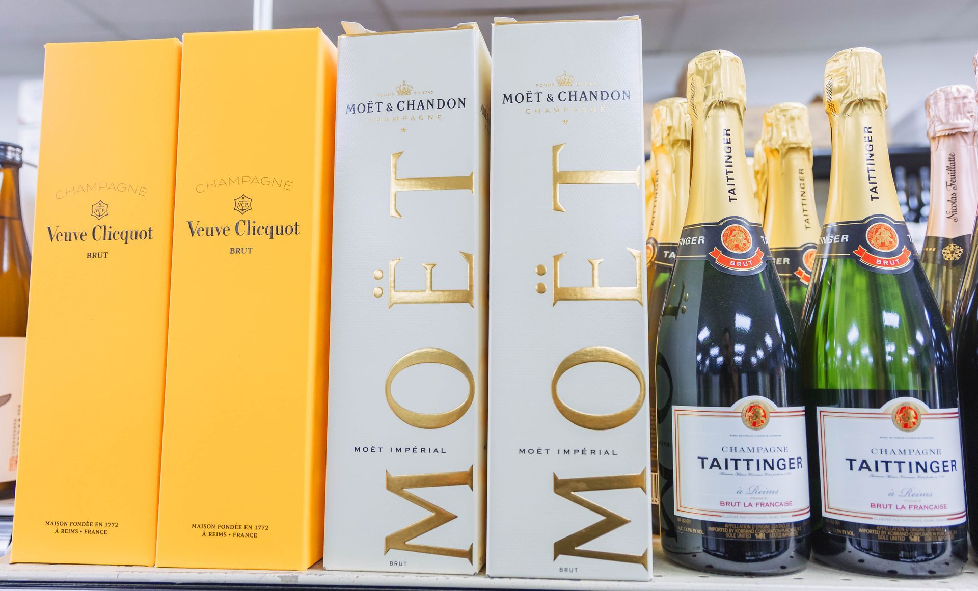 Champagne bottles and boxes on a shelf: Veuve Clicquot, Moët, and Taittinger, gold and white labels.