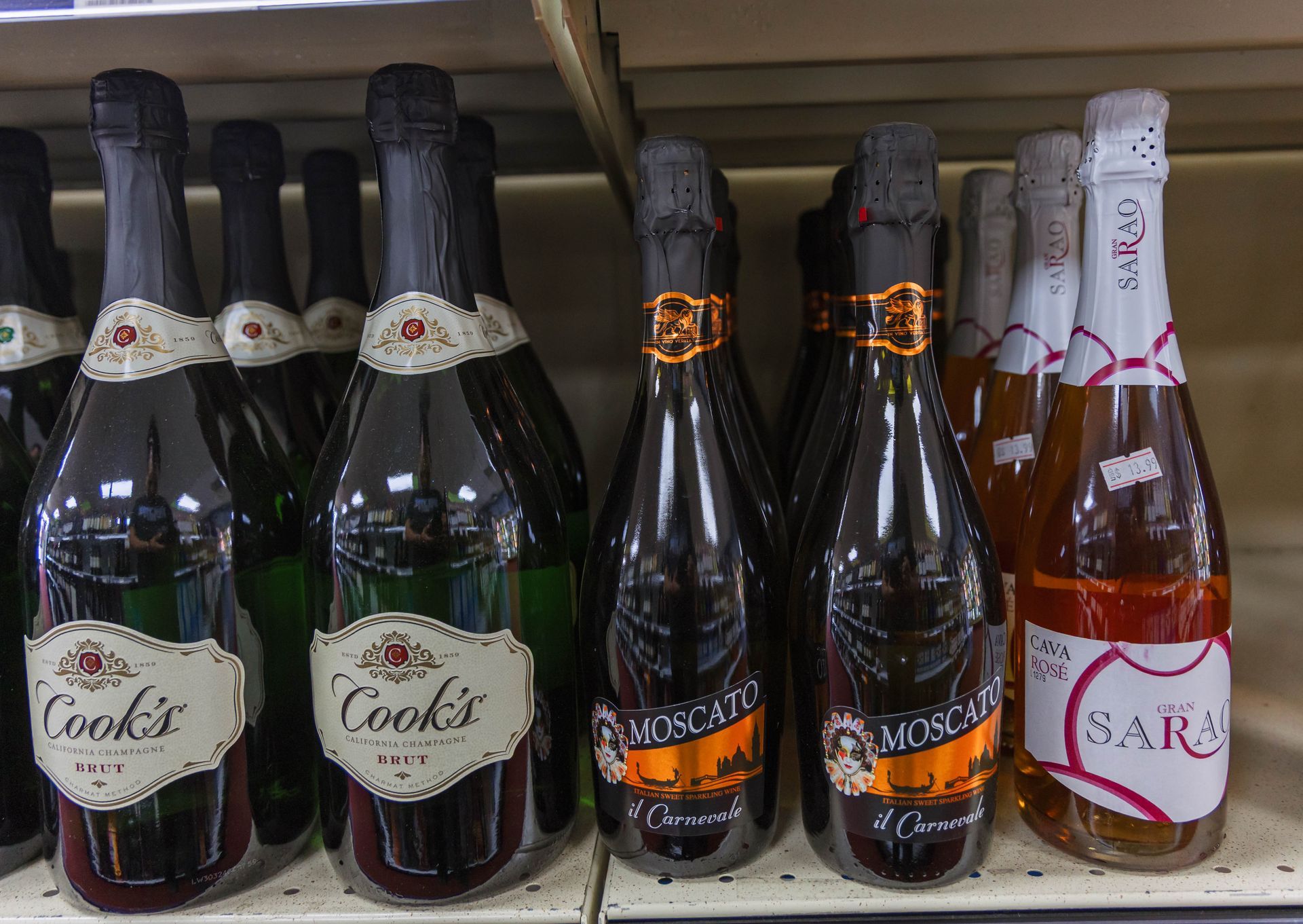 Bottles of sparkling wine on a shelf. Brands include Cook's, Moscato, and Sara. Dark bottles with foil and labels.