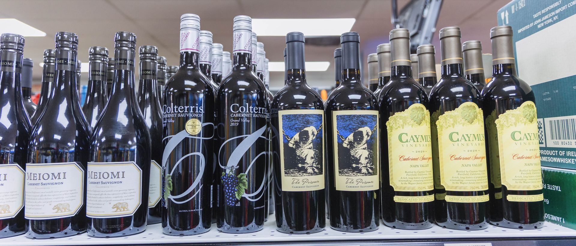 Wine bottles on a shelf in a store. Labels vary in color and design; mostly dark red bottles.