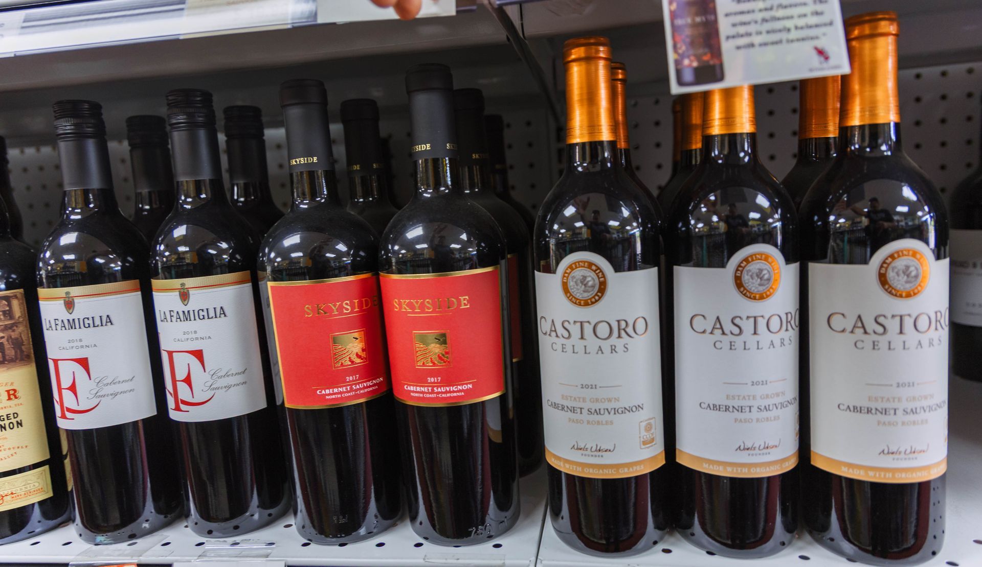 Bottles of red wine on a store shelf. Labels vary; gold and white labels, dark glass bottles, and bright red labels.