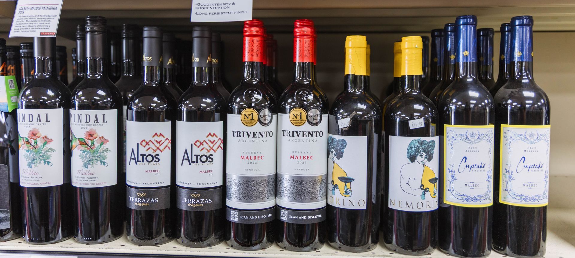 Bottles of red wine on a shelf, featuring colorful labels and foil tops in a shop.