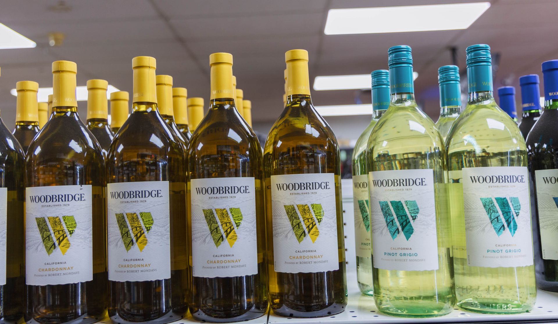 Bottles of white wine on a store shelf, yellow and blue caps, white labels with green logo.