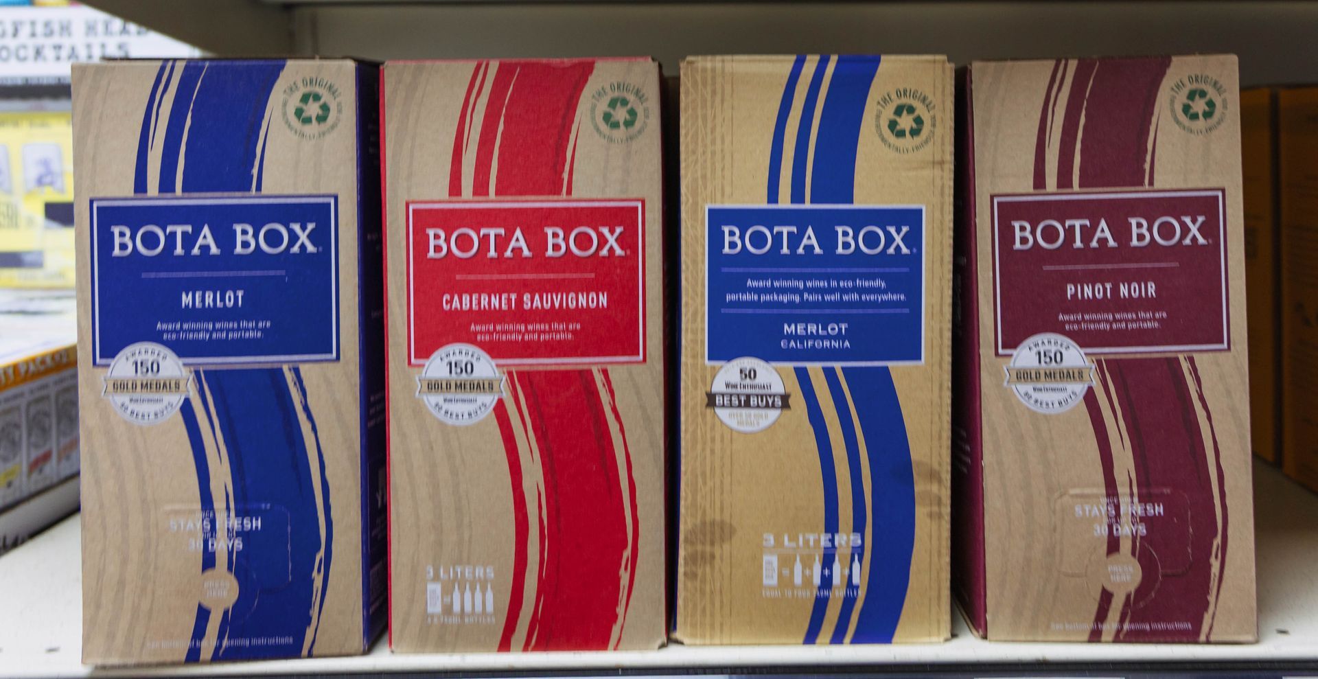 Four Bota Box wine boxes on a shelf; white, red, blue, and maroon labels with curved stripes.