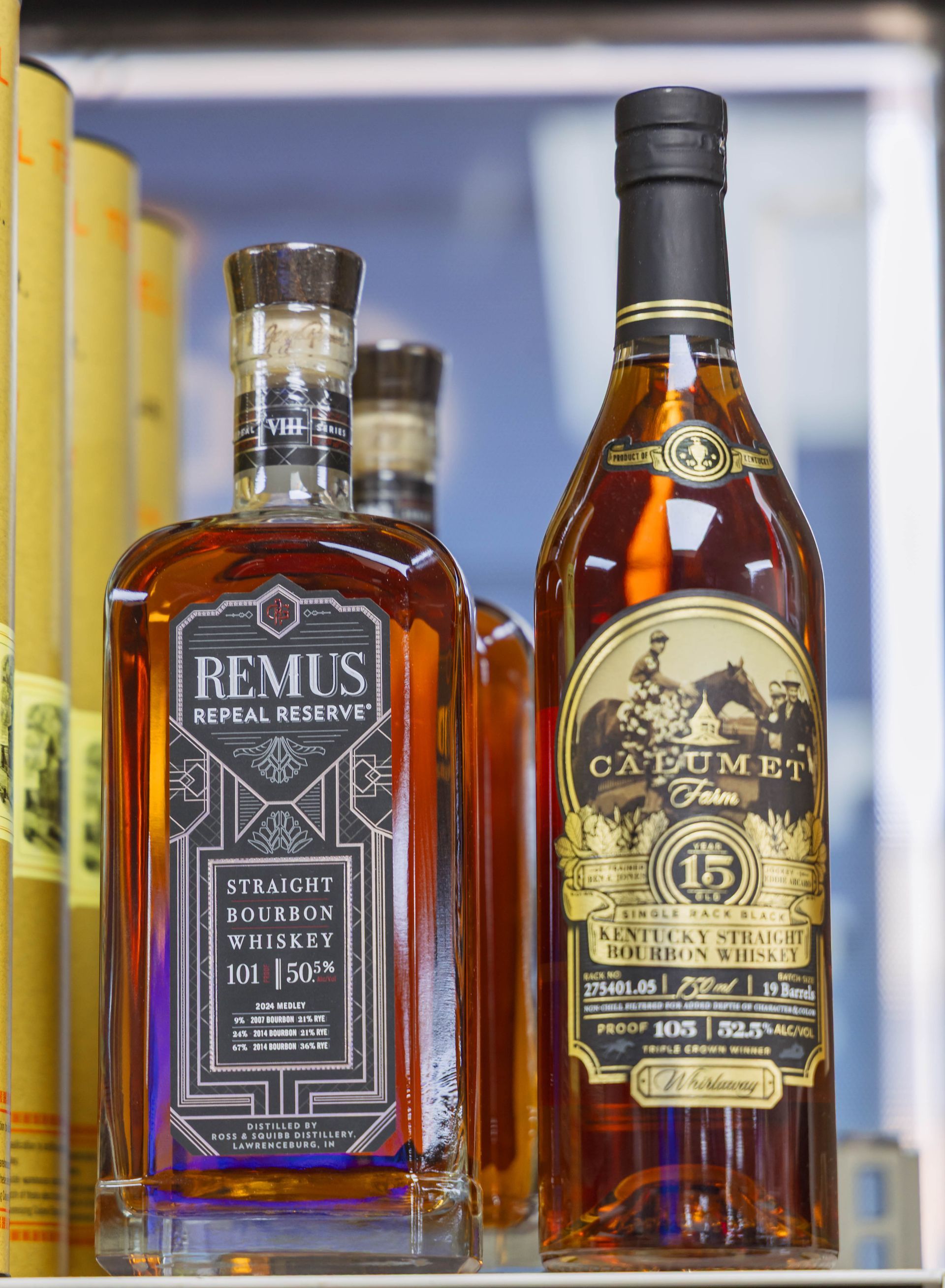 Two bottles of whiskey, Remus and Calumet, sit on a shelf with additional bottles in the background.