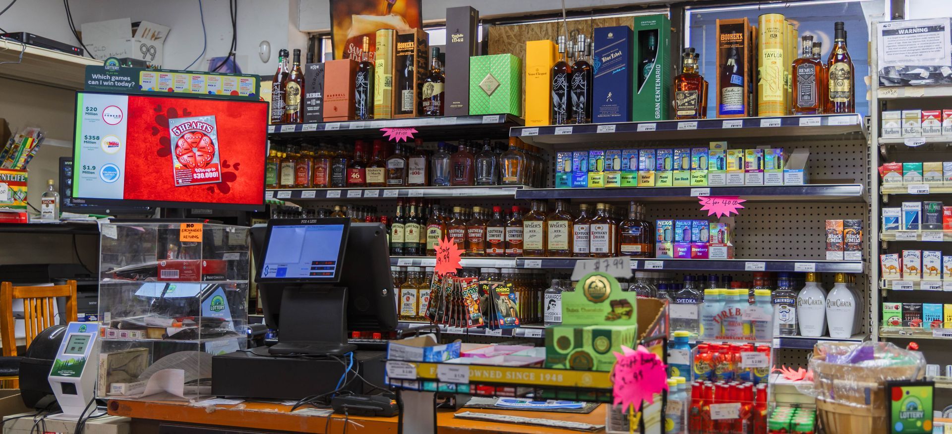 Interior view of a liquor store with shelves of bottles, a cash register, and a TV screen.