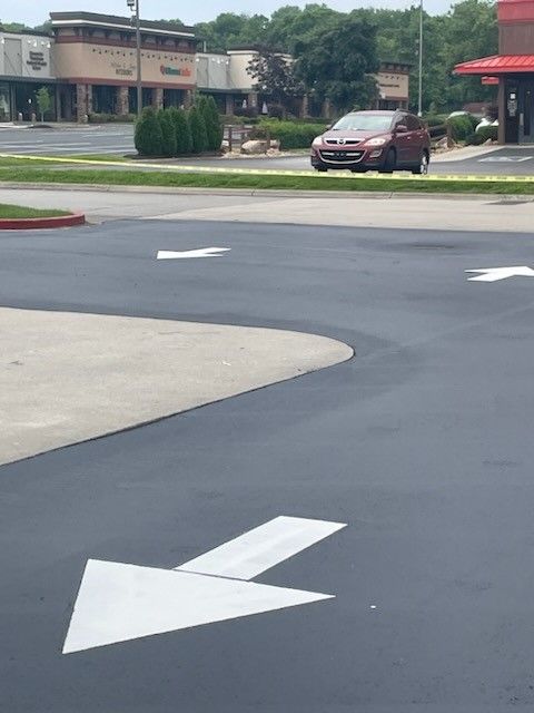 Asphalt road with white directional arrows, car driving; strip mall in background.