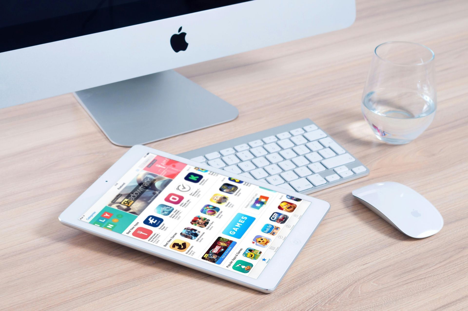 Tablet displaying app store, on a desk with computer, keyboard, mouse, and glass of water.