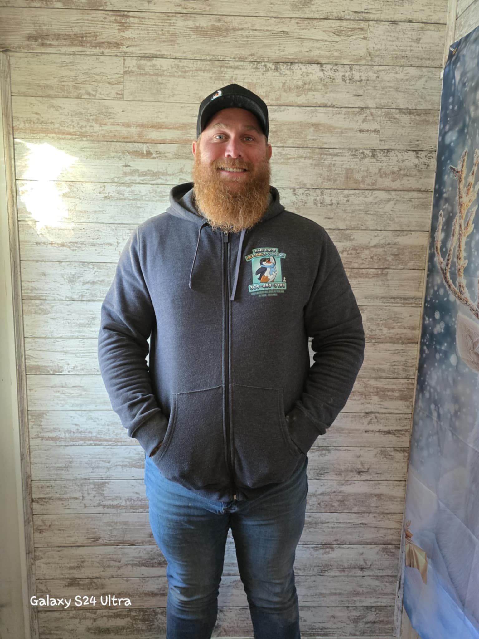A man with a beard is wearing a hoodie and standing in front of a wooden wall.