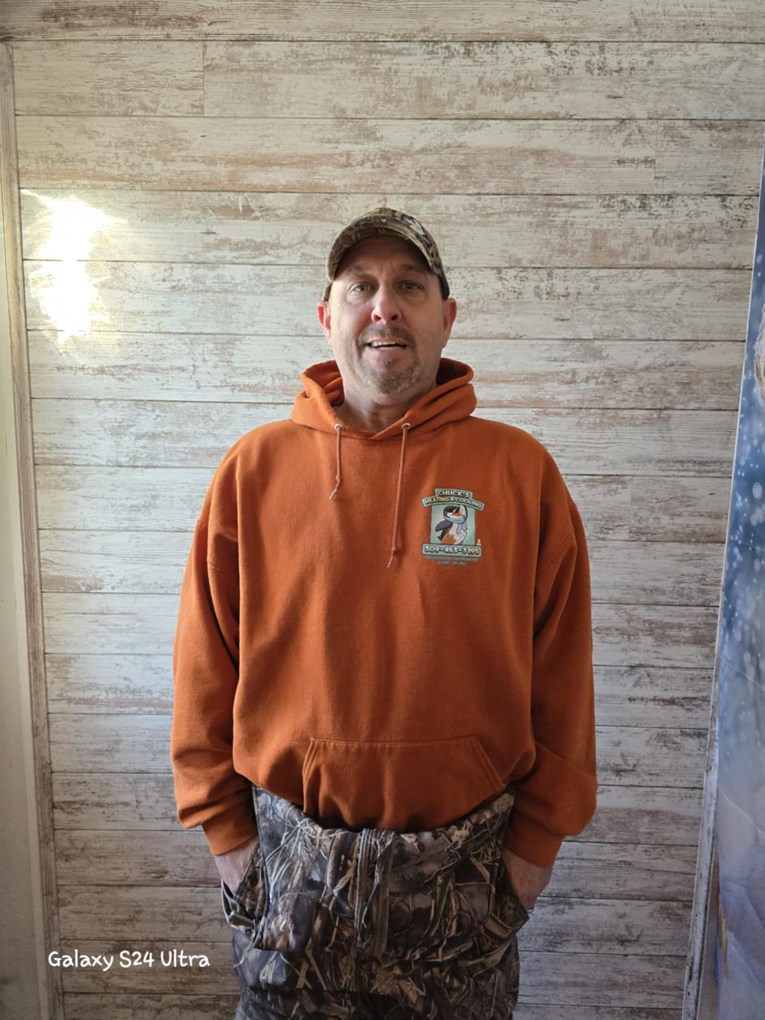 A man wearing a hoodie and camo pants is standing in front of a wooden wall.