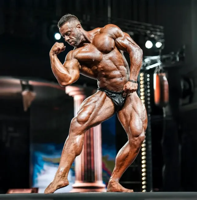 Bodybuilder flexing onstage, showcasing large muscles. Bronze skin, flexing pose, dark trunks, stage lights.